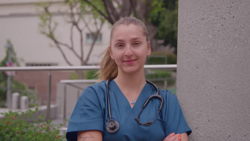 A young woman wearing medical scrubs and a stethoscope standing outdoors turns to be facing forward and smiles warmly