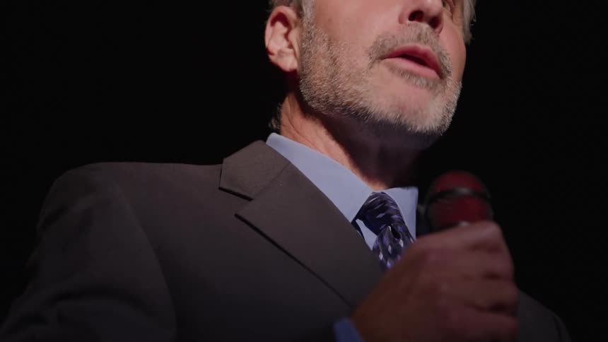 Graybearded man speaks into microphone in dark formal setting