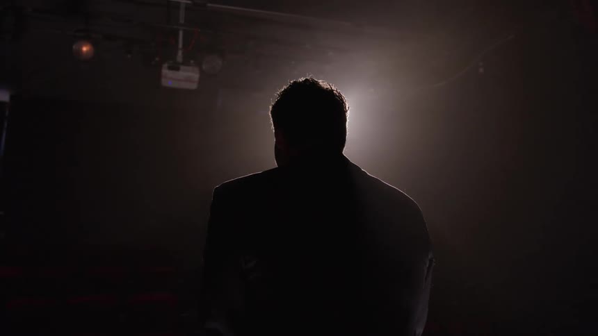 A person silhouetted from behind by a bright stage light stands in a dark room facing away with a projector visible near the ceiling