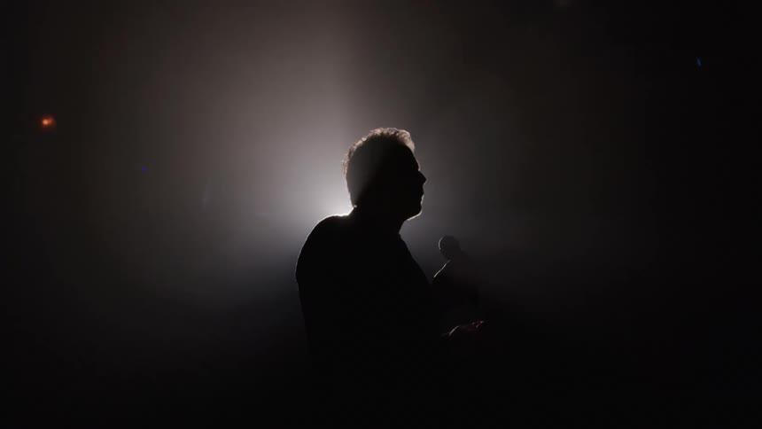 A man is captured in a medium closeup silhouette holding a microphone while standing on a stage dramatically outlined by a brilliant white light source behind him in a hazy dark environment
