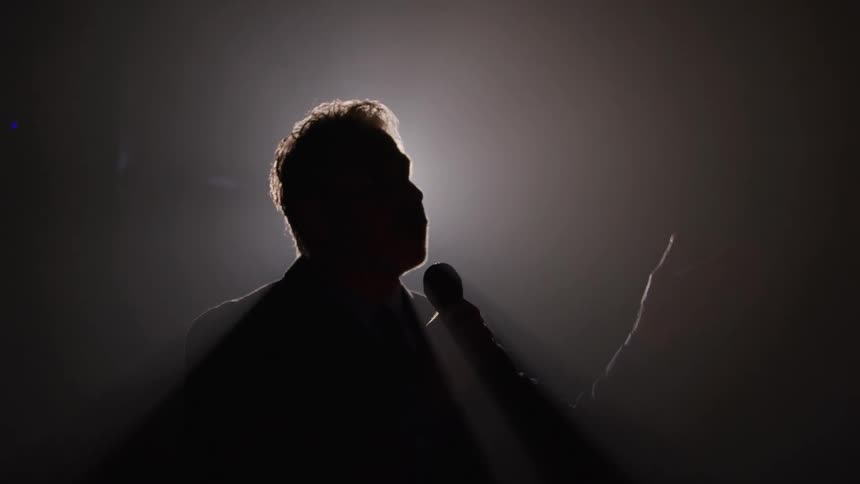 A person dramatically silhouetted in profile holds a microphone and gestures with their free hand