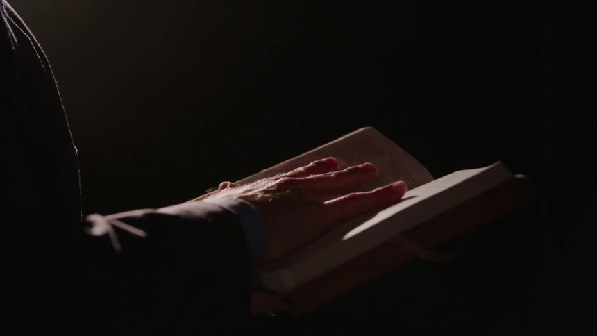 In a dark setting a man holds an open book and gestures dramatically over the pages