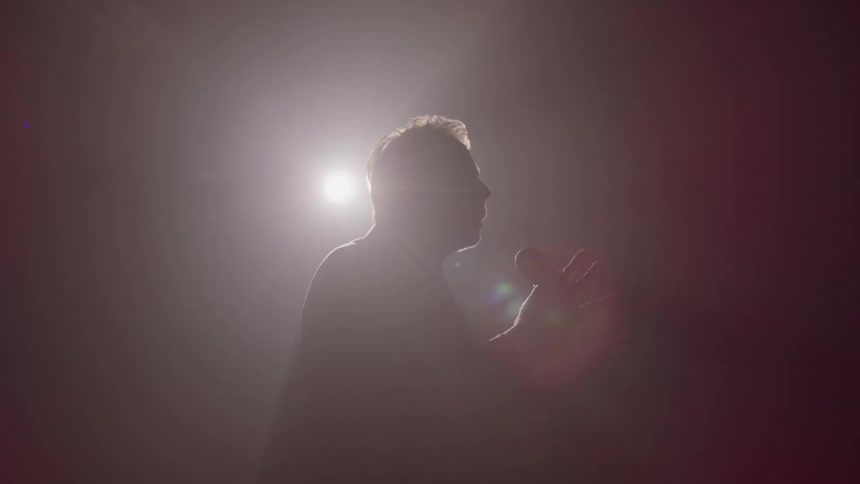 A person silhouetted against a bright background light is seen speaking into a microphone while gesturing with their hands