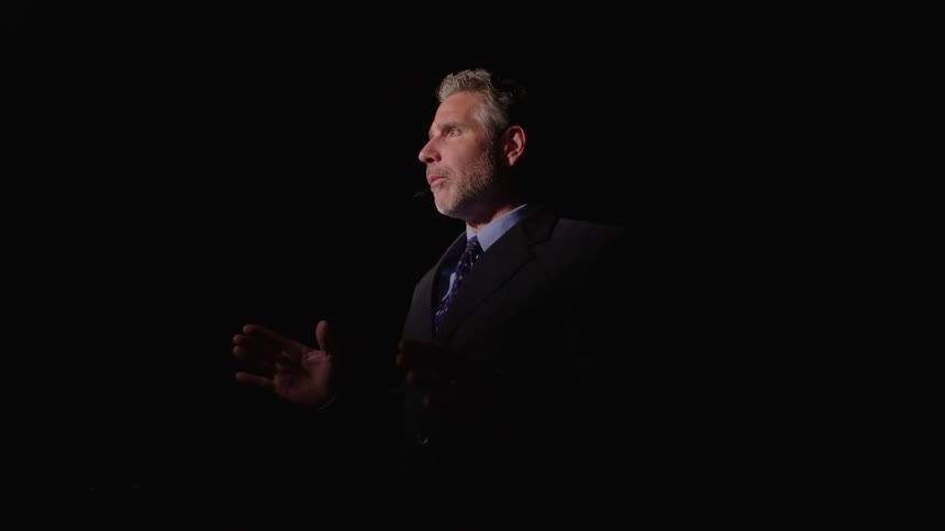 Man in suit speaks with expressive gestures under side lighting in dark setting