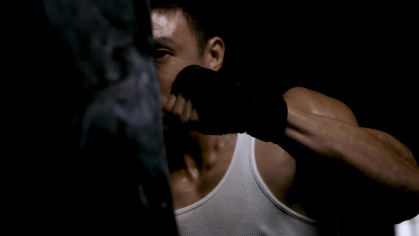 A muscular sweaty man wearing a tank top and hand wraps trains intensely by repeatedly punching a dark punching bag in a dimly lit setting