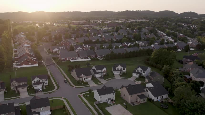 Aerial view of neighborhood houses and cars in driveways and on the street as the sun glows in the distance