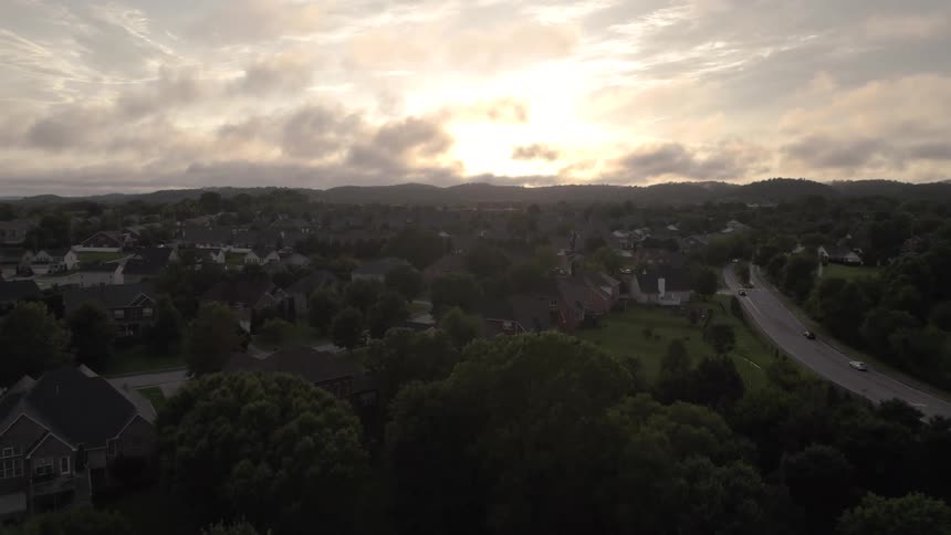 Aerial view of neighborhood houses and trees with traffic on the street as the sun shines through clouds on the horizon