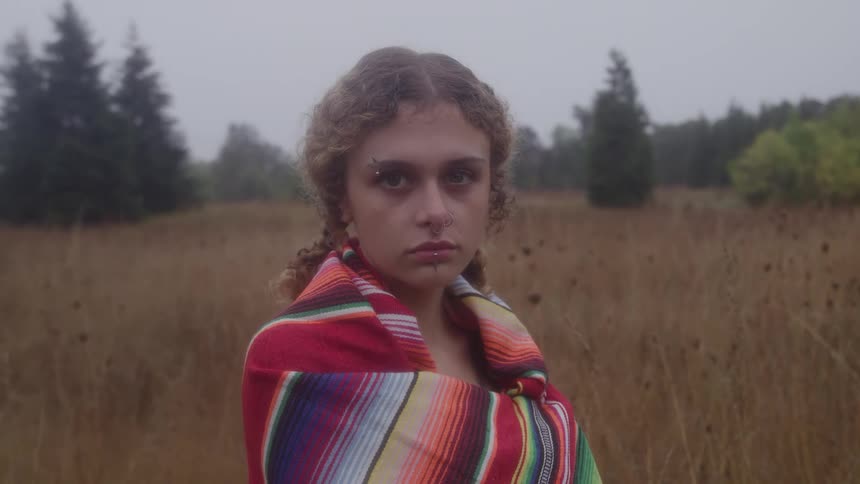A teenage girl with light curly hair and facial piercings wrapped in a bright multistriped blanket turns slowly from a profile view to stare intently into the camera while standing in an overcast open field of dry tall grass