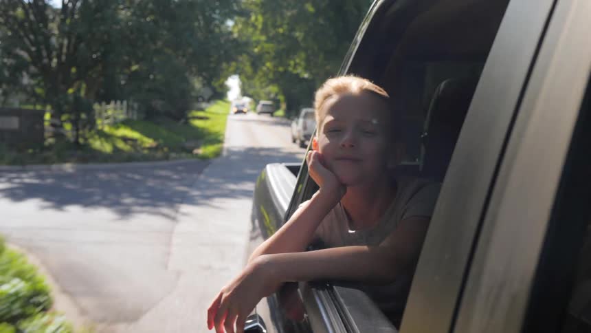 A child with blonde hair leans out of a moving vehicles rear window resting their chin on their hand and smiling faintly while traveling down a sunny treelined road