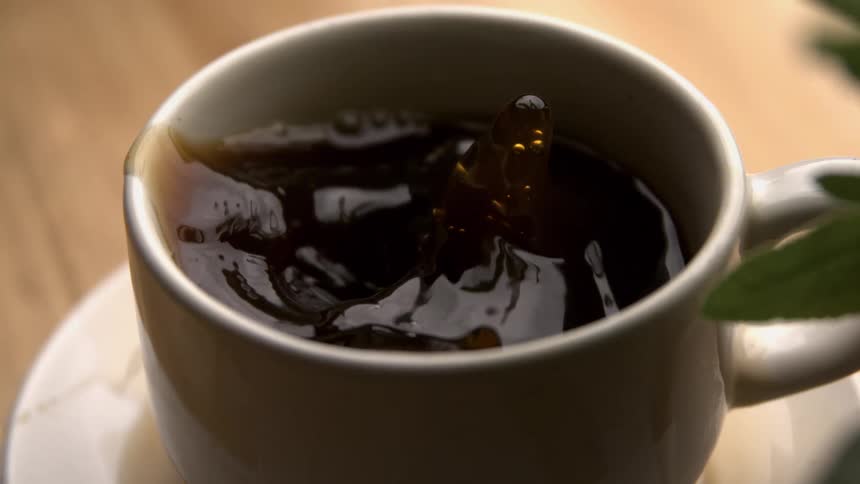 A closeup shot focuses directly into a white cup and saucer filled with a dark liquid where drops of the same dark liquid are falling causing the surface to ripple and splash outward