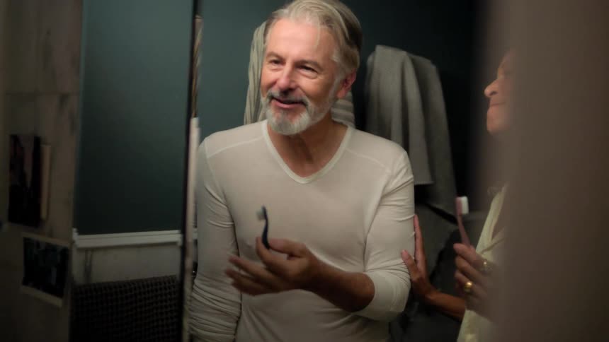 A happy couple brushes their teeth together at a sink in front of a well lit mirror in a bathroom