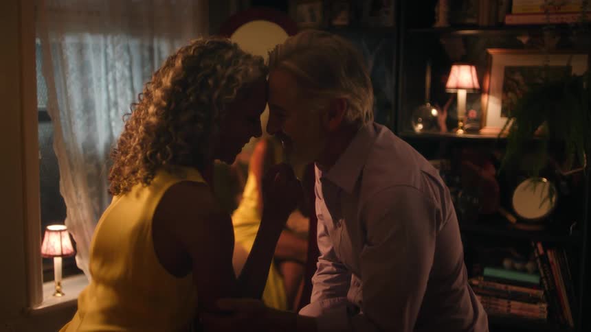 A man and a woman are standing close together in a dimly lit richly decorated room sharing an intimate kiss while the womans reflection is simultaneously visible in a large oval mirror behind them