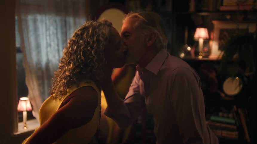 A man and a woman are seen in a close embrace sharing an intimate kiss in a dimly lit cozy room adorned with bookshelves and lamps as the womans seated reflection is visible in a large central mirror
