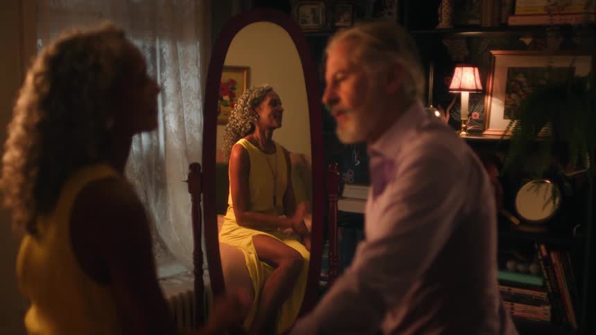 A man is talking to a woman who is sitting on a stool with a reflection of her image in the mirror behind her