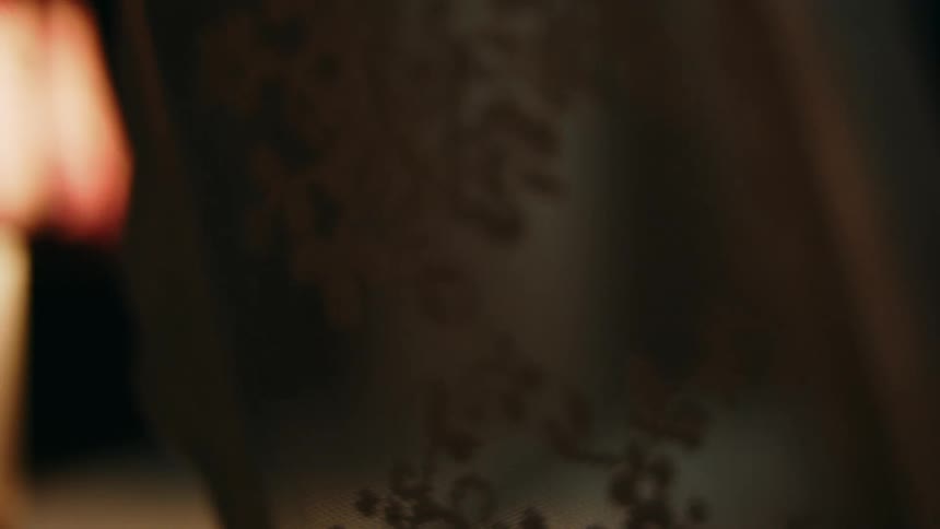 Soft amber light grazes the delicate floral patterns of a lace fabric highlighting the intricate texture as it shifts gently against the surrounding darkness