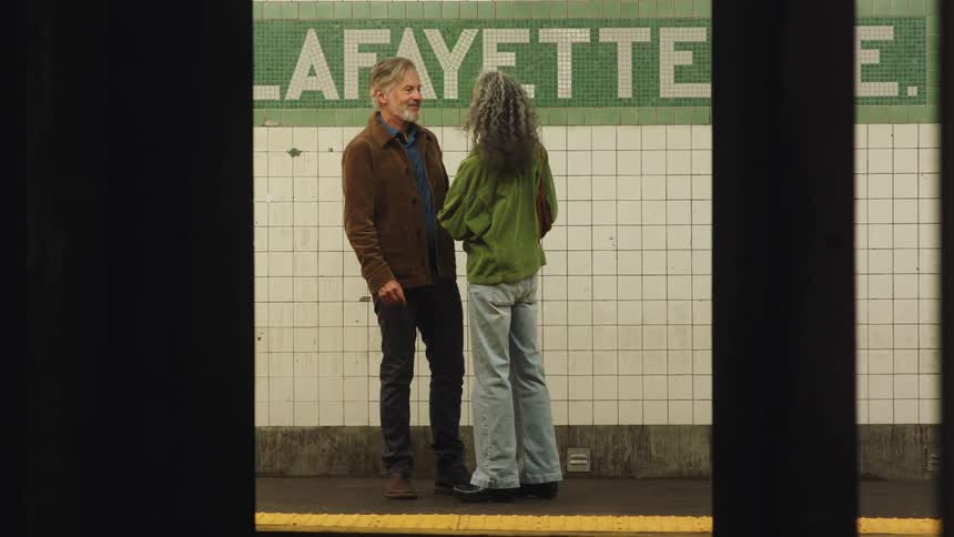 A stylish looking older couple are standing on a subway platform smiling and hugging as a train travels by