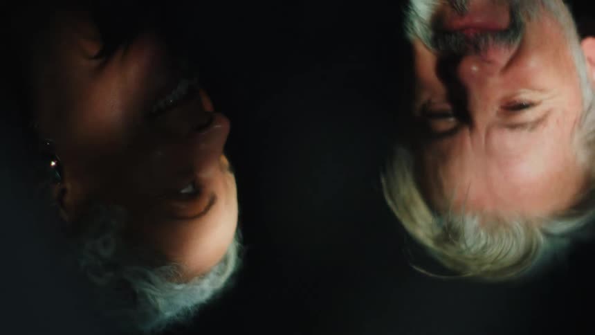 A reflection of an older man and woman with gray hair illuminated by bright dramatic lighting that contrasts sharply with the black background as they gaze toward each other