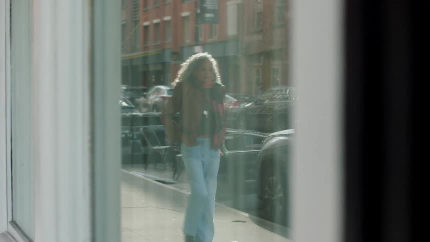 A person with light curly hair dressed in a jacket and light jeans walks quickly past the large reflective window of a storefront with their figure briefly visible in the glass reflection of a busy redbrick urban street scene