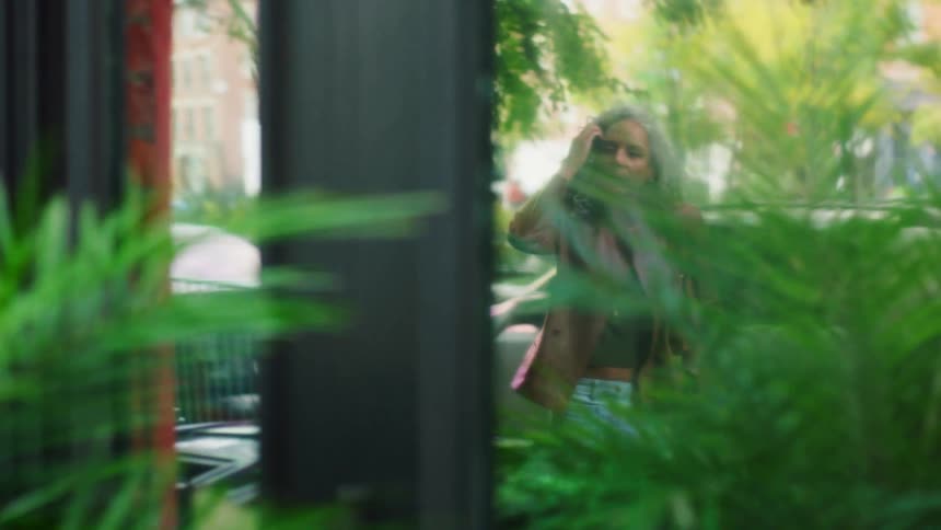 A woman briefly walks into the frame seen through a large glass window and partially obscured by vibrant green indoor or outdoor plants she is wearing a brown jacket and jeans and the background features an orange brick wall and a blurry street scene