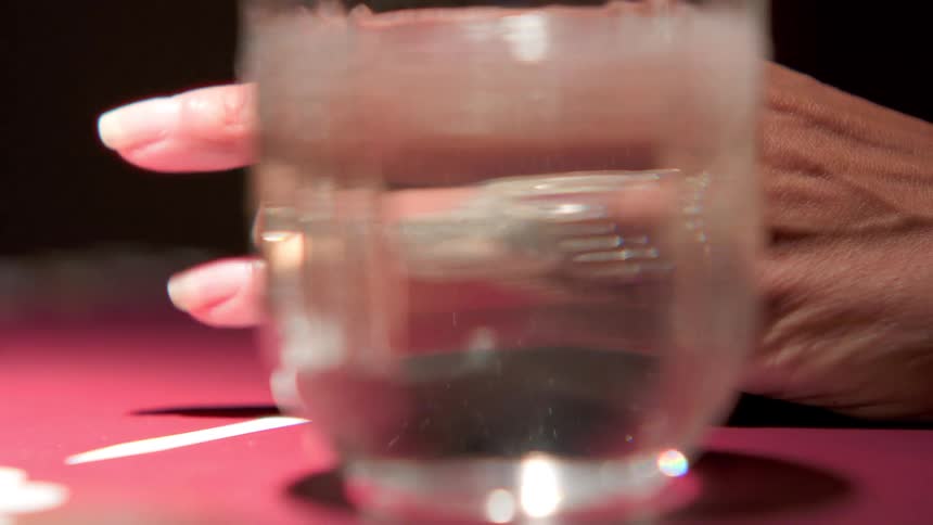 A hand wearing a diamond ring is reflected through a clear glass of water on the table