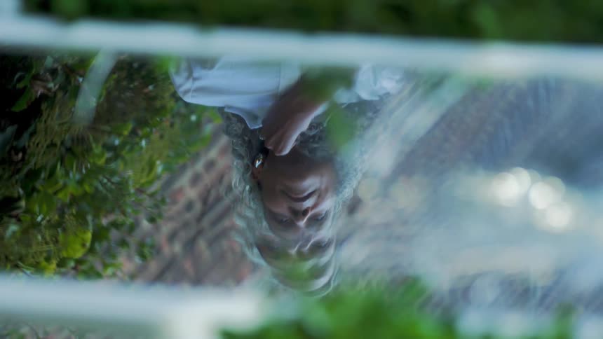 A closeup shot captures the inverted and distorted reflection of an older persons face wearing a white shirt and touching the surface which is surrounded by the vibrant dark green reflections of an outdoor environment