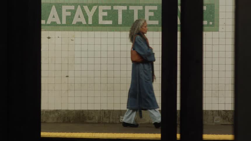 The video framed through the dark vertical bars of a train door shows a woman with long gray hair walking along a subway platform the platform wall is covered in white square tiles with a green border that displays the word lafayette