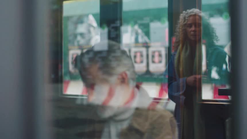 An older man with gray hair and a beard wearing a brown jacket sits indoors working on something viewed through a reflective window the window catches the blurry reflection of a woman with curly hair standing outside briefly overlapping the man in th