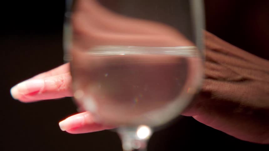 A closeup shot focusing on a hand holding a wine glass filled with a clear liquid likely water the hand has long wellmanicured fingernails with a natural pinkish polish the glass is slightly out of focus causing a refraction and distortion