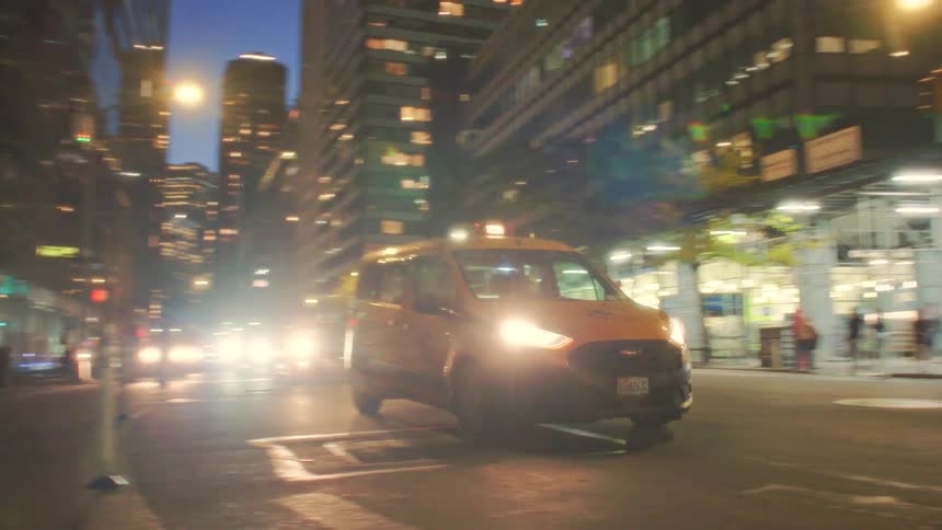 A yellow taxi van drives quickly across a welllit city street at nightthe surrounding skyscrapers reflect the bright glare of headlights and streetlights creating a dynamic urban scene