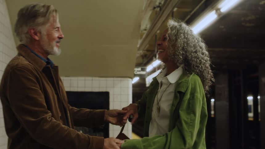 An older couple joyfully dances together on a subway platform while waiting for the train