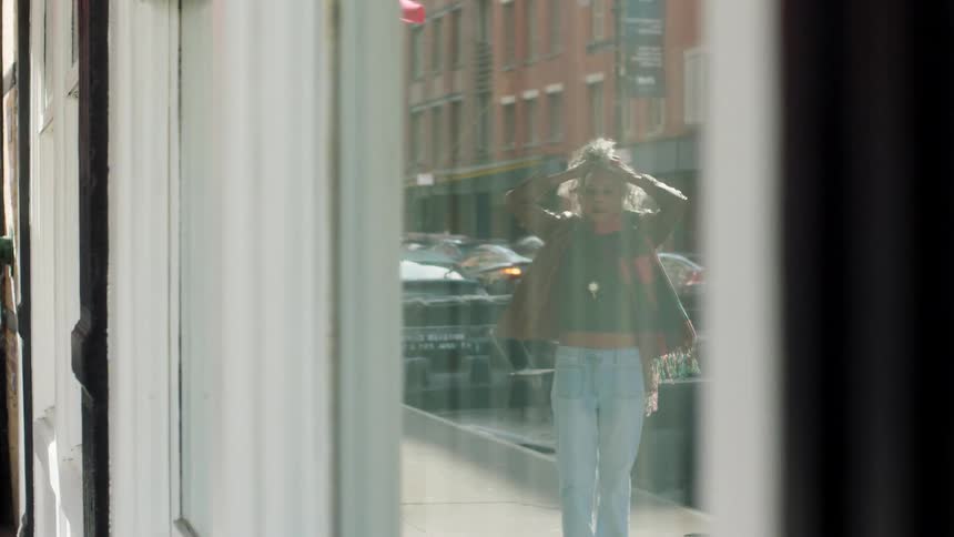 A young person with bright curly hair walks past a storefront their reflection briefly captured in the large window the background street view shows city buildings and parked cars suggesting a busy urban area