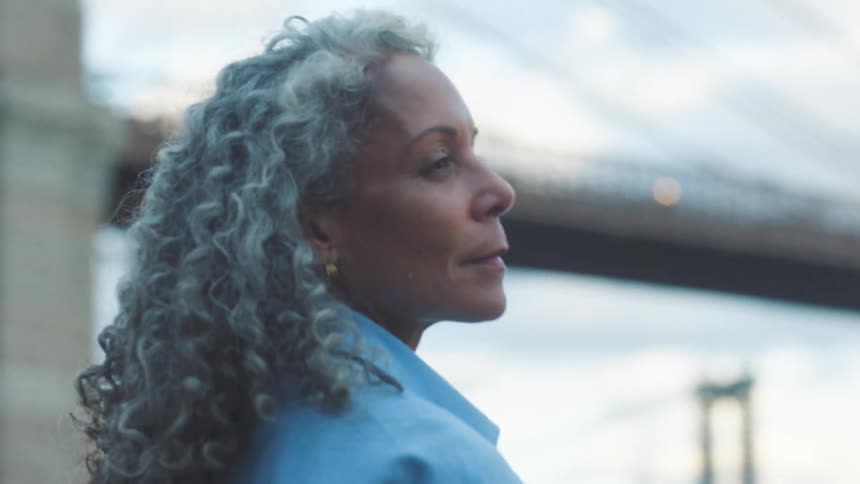 A middleaged woman with grey curly hair stands on a riverbank at midday a bridge visible in the background she looks emotional yet carries a gentle smile on her face