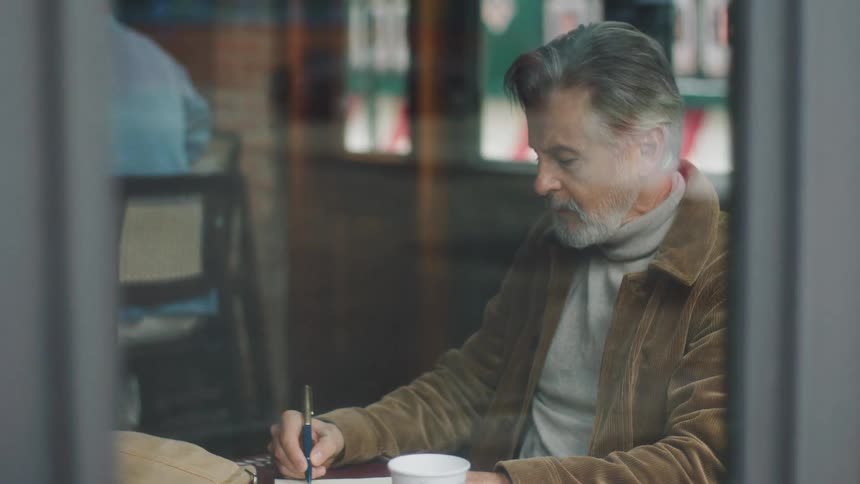 A middleaged man sits inside a caf at midday a cup of coffee placed in front of him wearing a brown jacket his grey facial hair visible he writes with a pen in a notebook