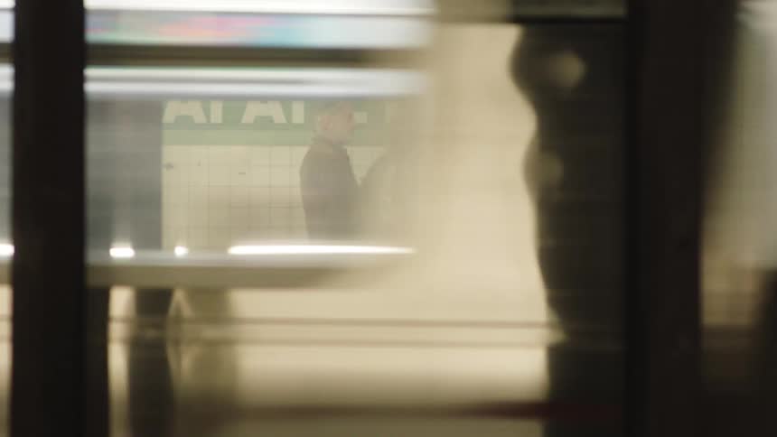 A middleaged woman and a man embrace in the train terminal as a train drives by in the background