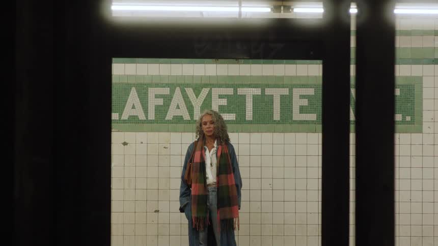 A middleaged woman with grey curly hair stands in a train terminal at midday wearing a jacket with a scarf draped over her shoulder she watches a train drive by before turning and walking out of the terminal