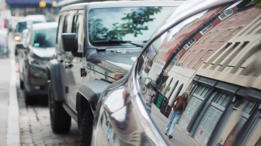 A dark jeep wrangler is parked on an urban street lined with cobblestones and a few other vehicles with a highly reflective side surface of the car in the foreground showing a distorted but clear reflection of a female walking by brick buildings