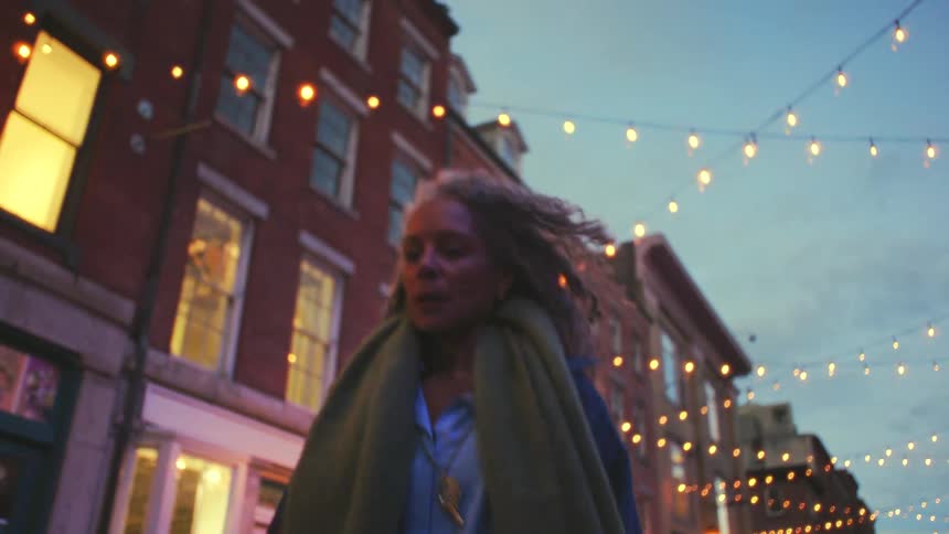 An older woman walks along a city street lined with buildings and string lights at dusk