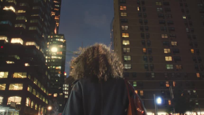 Person in denim jacket walking at night among lit buildings in busy urban setting
