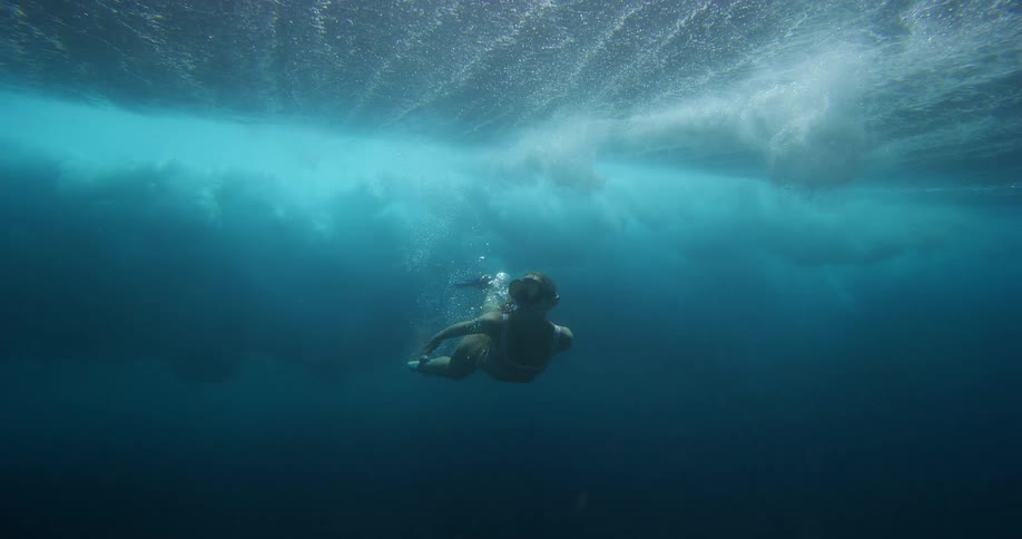 A person wearing flippers dives downward into deep clear blue water viewed from below the surfacethey swim toward the light filtering down from the agitated water surface above leaving a trail of bubbles