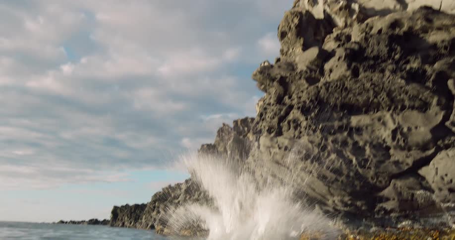 A young man leaps backward from a jagged cliff spinning through the air against a cloudy blue sky before splashing heavily into the sea