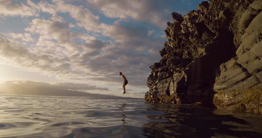 A young man jumps off a cliff into the ocean at sunset