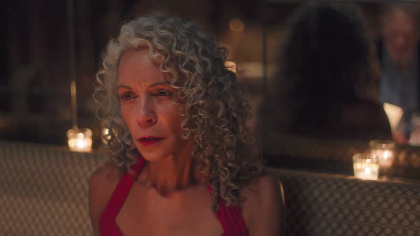 The video opens with an outoffocus shot of a woman with gray curly hair sitting in a dimly lit restaurant booth with a man seated across the tablethe camera then focuses closely on the womans face as she appears to be speaking passionately or emo