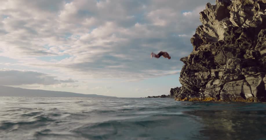 A person flips mid air off a tall dark cliff into the ocean under a partly cloudy sky