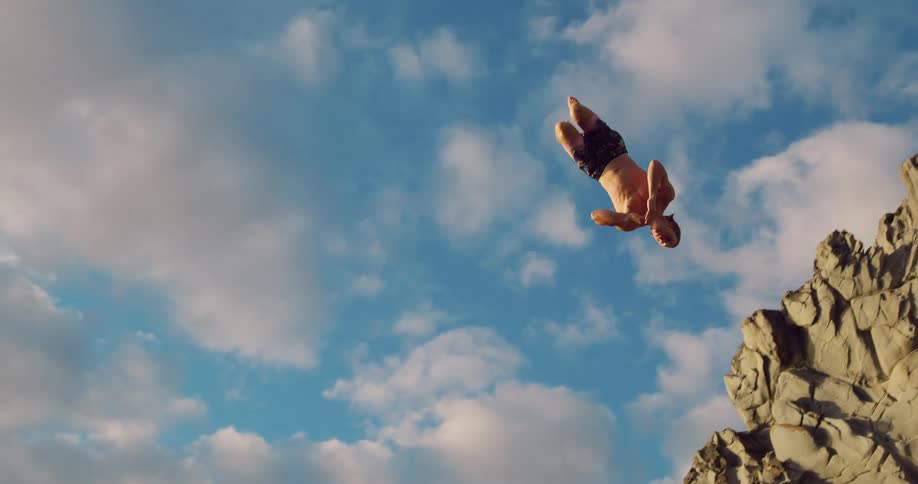 Person leaps from the top of the rocks toward the sea under a blue sky with clouds