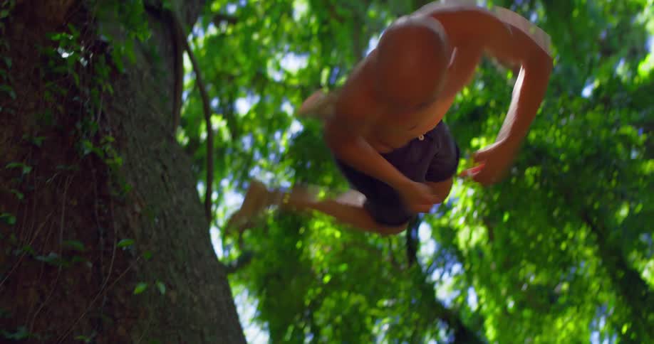 A topless male runs up a large tree trunk and does a backwards flip in a forest area