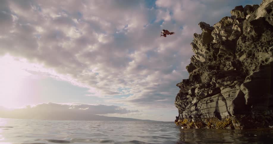 A man jumps from a tall rugged dark cliff face that juts out over the calm ocean water all set against a dramatic sky with scattered clouds and the bright glow of the sun near the horizon