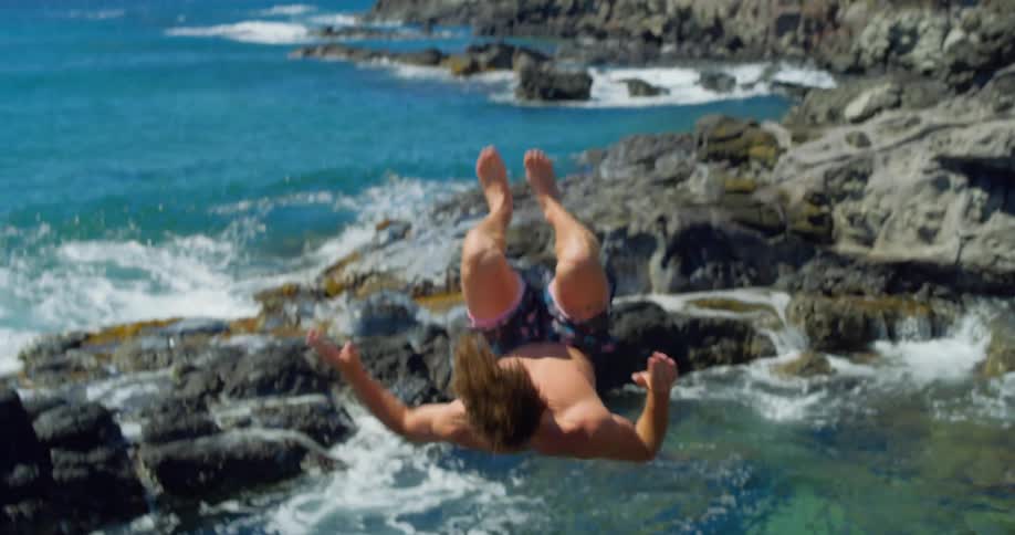 A person jumps off rocky coastal cliffs toward the ocean under a bright sky