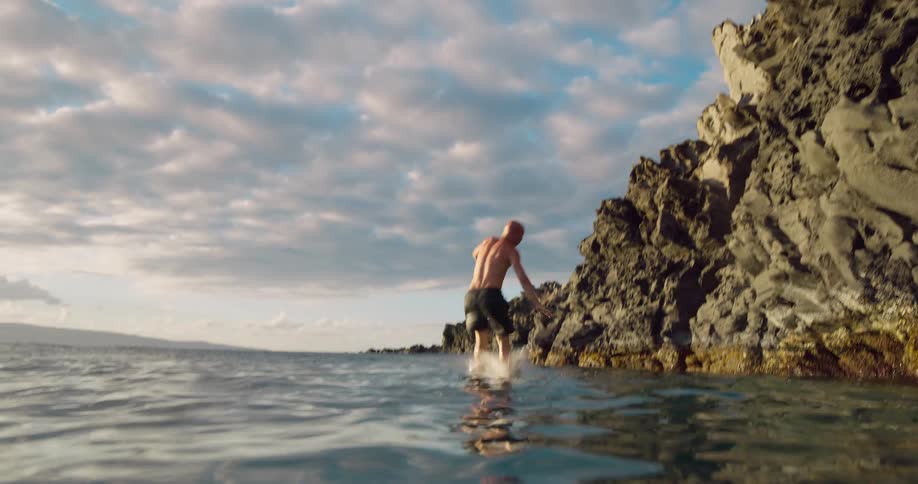 Person performs a backflip from the rocks toward the sea under a cloudy sky