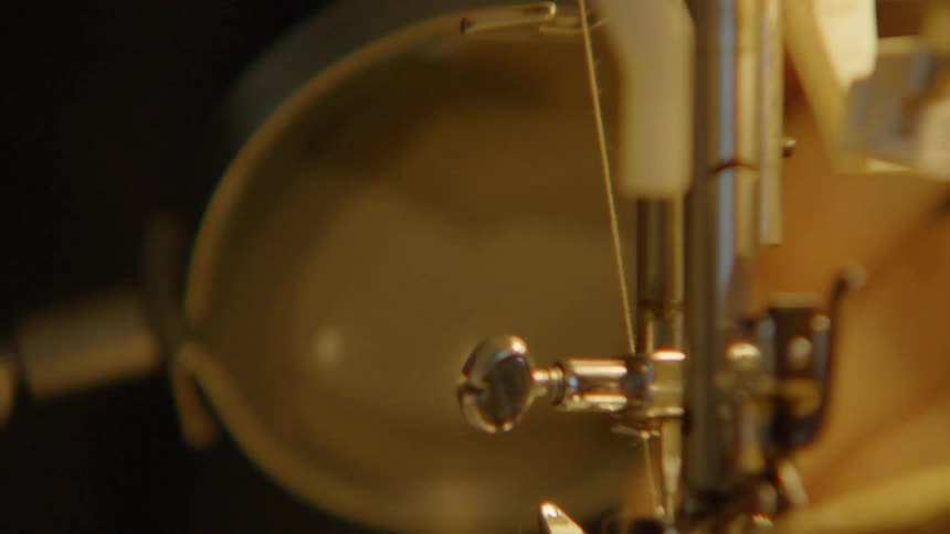 A persons mouth and hands are near the bright light of a white sewing machine guiding a piece of striped tan fabric under the needle and presser foot while an auxiliary magnifying attachment is visible on the left