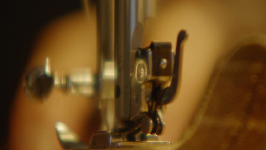 A very tight closeup focuses directly on the chrome sewing machine needle and the metal presser foot as they rest on a piece of tan fabric as a hand guides the fabric all rendered against a dark warmtoned and outoffocus background
